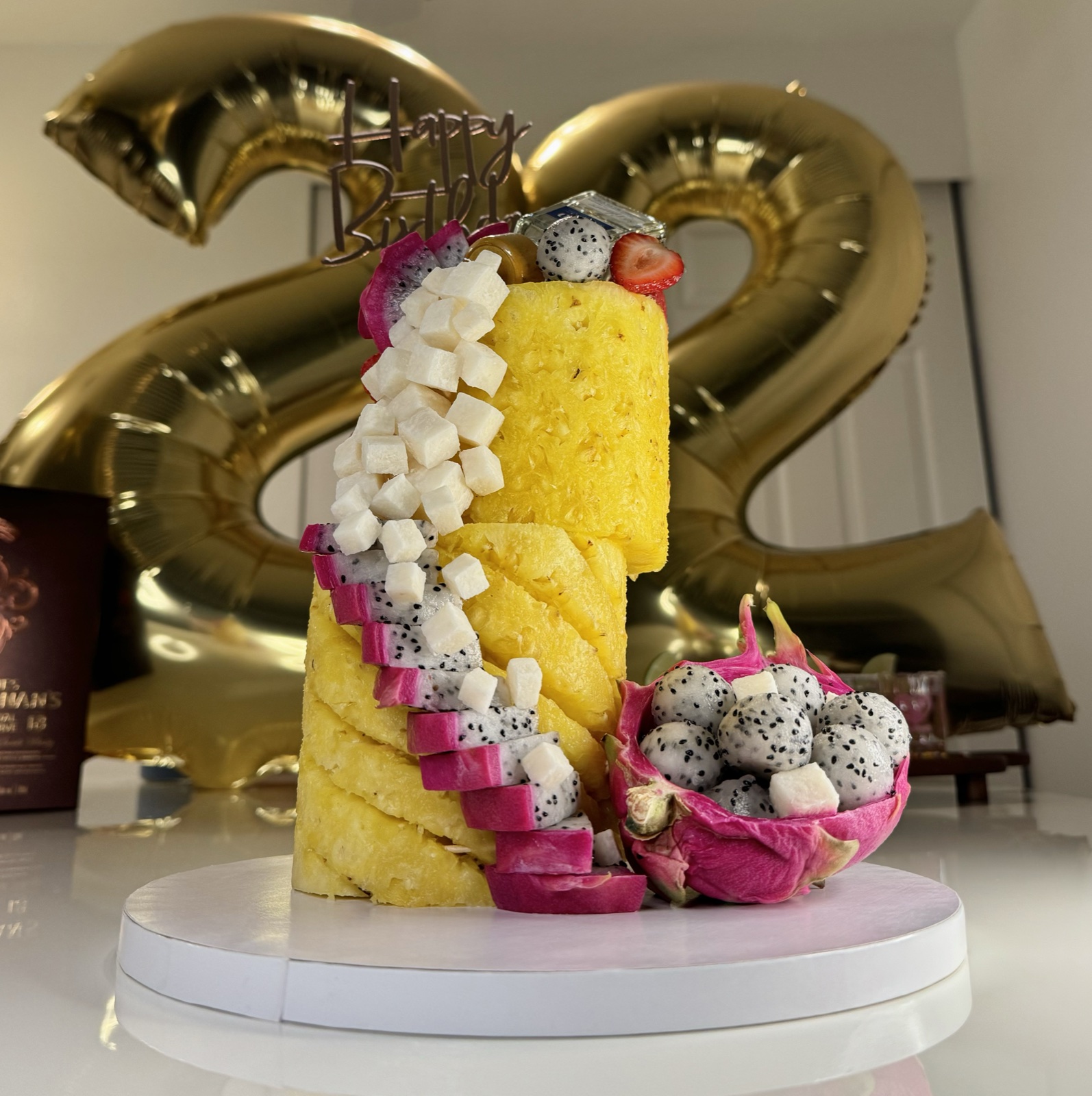 Two-tier pineapple cake with dragon fruit and jícama with a Happy Birthday topper and gold balloons